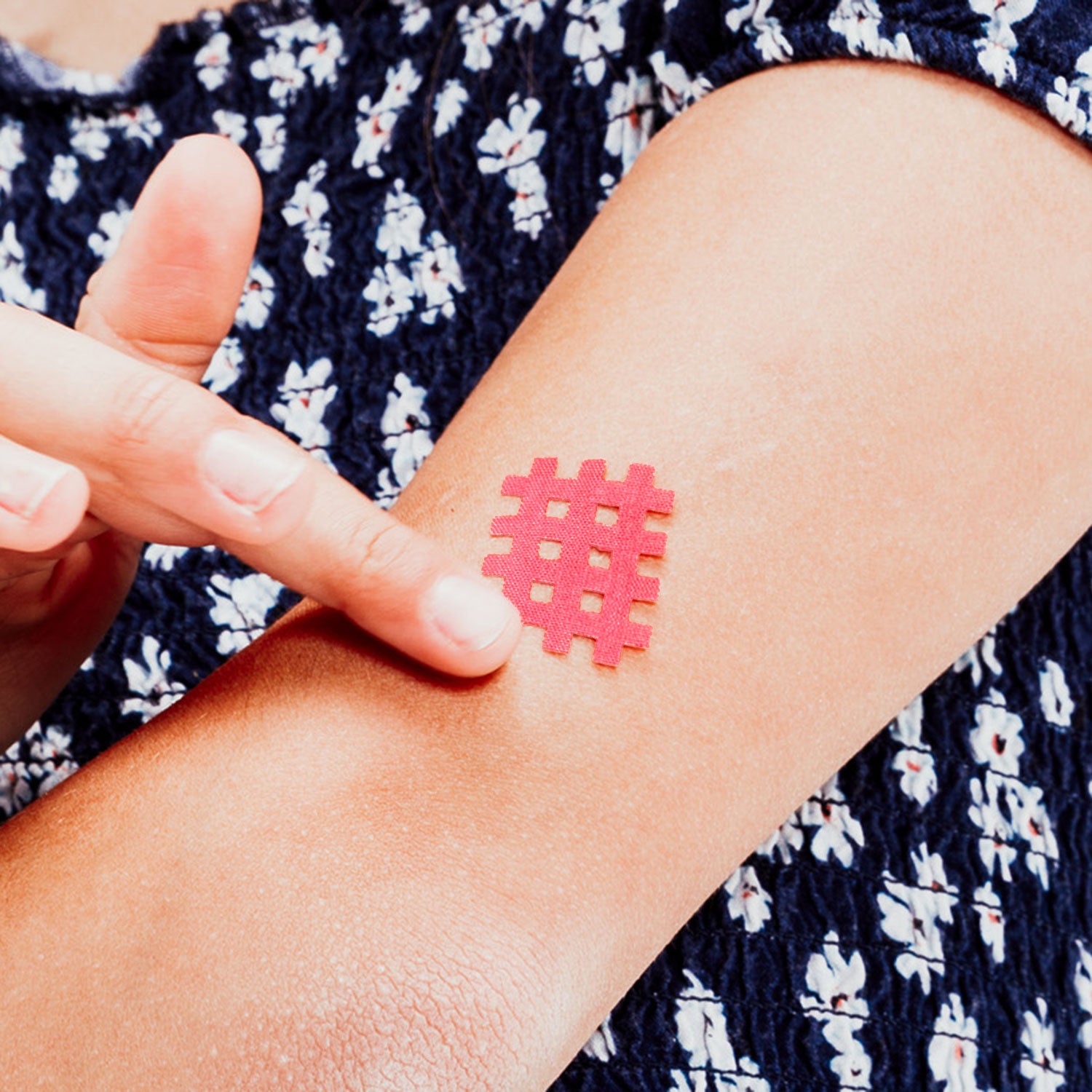 Finger berührt ein pinkfarbenes, gitterförmiges Moskinto-Pflaster, das auf dem Unterarm einer Person klebt. Die Person trägt ein dunkelblaues Oberteil mit weißem Blumenmuster.